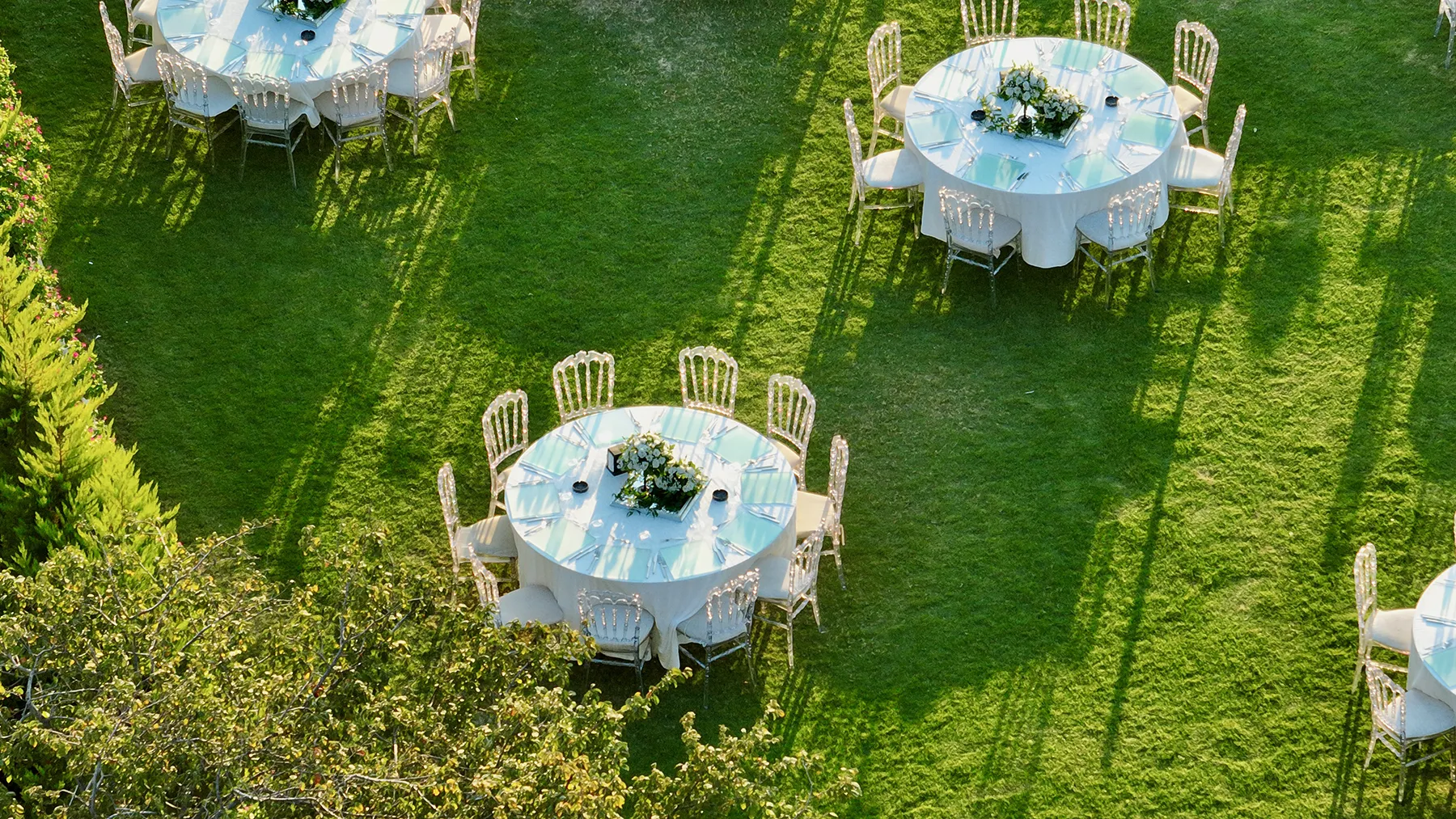 Outdoor garden event with round tables, white linens, and clear chairs arranged on a green lawn for an upscale private gathering