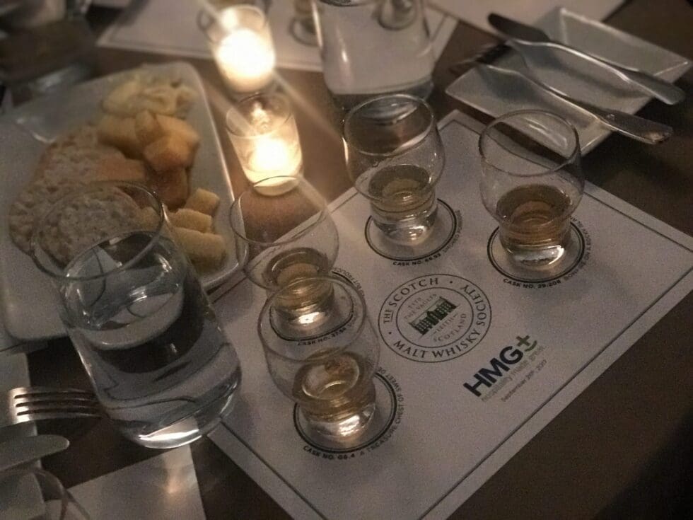 A dimly lit Scotch malt whisky tasting setup featuring several small glasses of whisky arranged on an HMG+ branded tasting mat from the Scotch Malt Whisky Society, accompanied by a water glass, bread and cheese, and a flickering candle.