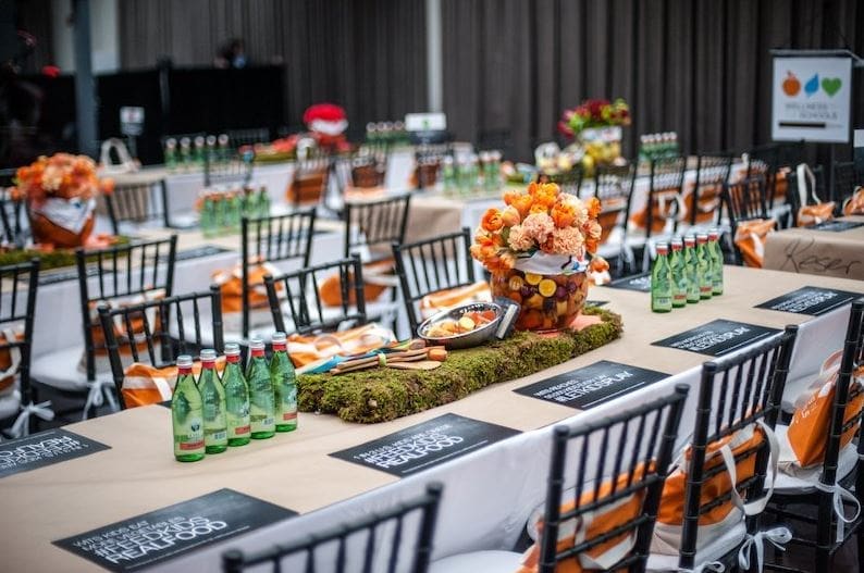 An elegantly styled event banquet hall with long tables set with black Chiavari chairs, green glass water bottles, black printed place cards, orange floral centerpieces atop moss-covered risers, and orange and white striped gift bags tied to chair backs.
