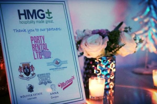 An HMG+ Hospitality Made Great event signage card reading 'Thank you to our partners,' listing sponsors including Party Rental Ltd., Anchor Distilling Company, Murray's, Wölffer Estate Vineyard, and Anna Coronel, displayed alongside a white rose floral arrangement and glowing candles in a warmly lit event setting.