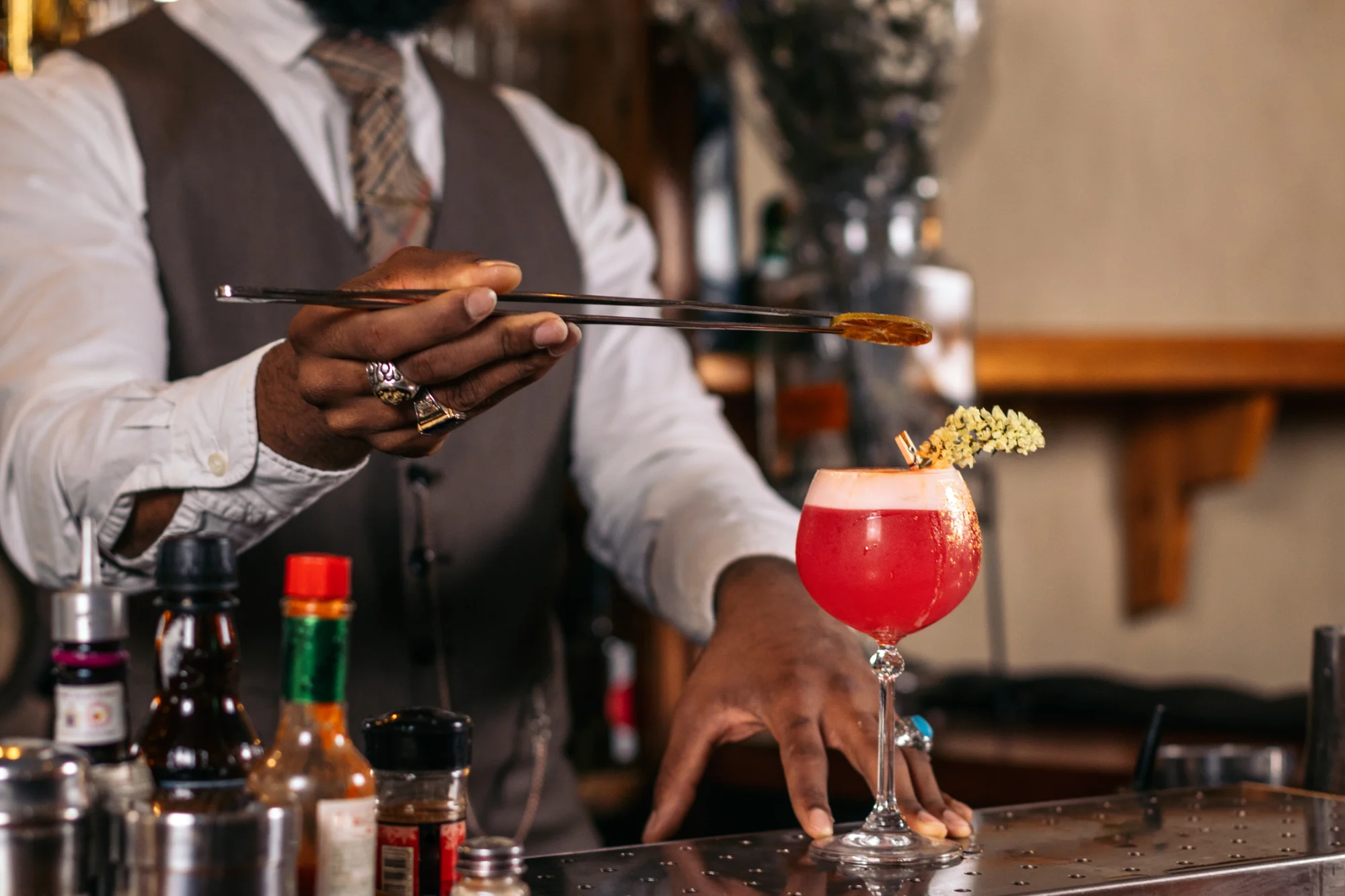 A bartender in a vest and tie uses tongs to garnish a red cocktail in a coupe glass with a dried citrus slice, surrounded by bottles on a bar counter.