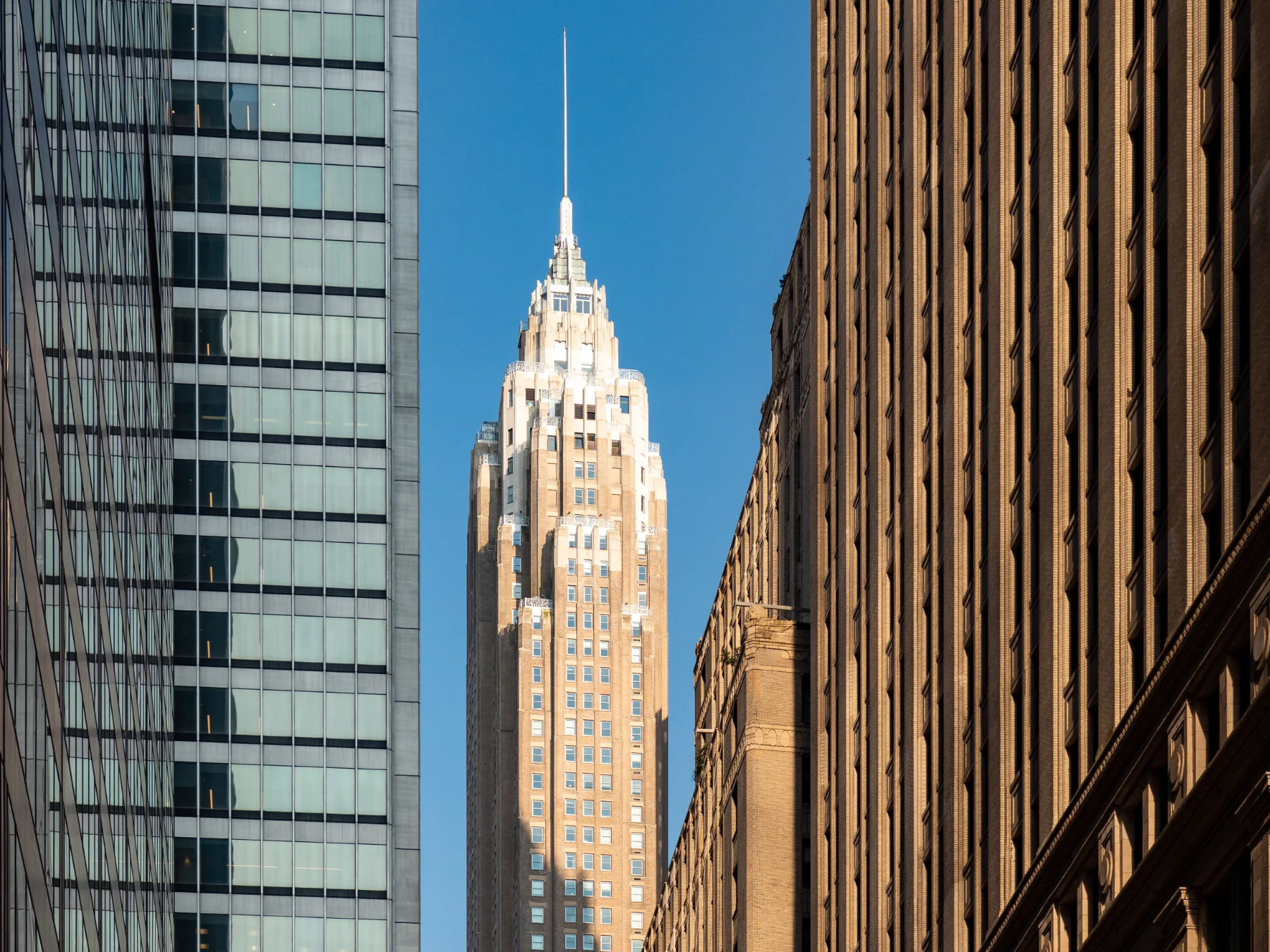 art-deco-skyscraper-spire-city-hmg-plus. Art deco skyscraper with a spire framed between two buildings in daylight